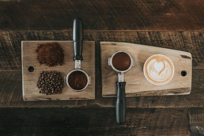 A photo of several coffee related items on a timber chopping board. It includes a pile of coffee beans, ground coffee, and a cappuccino