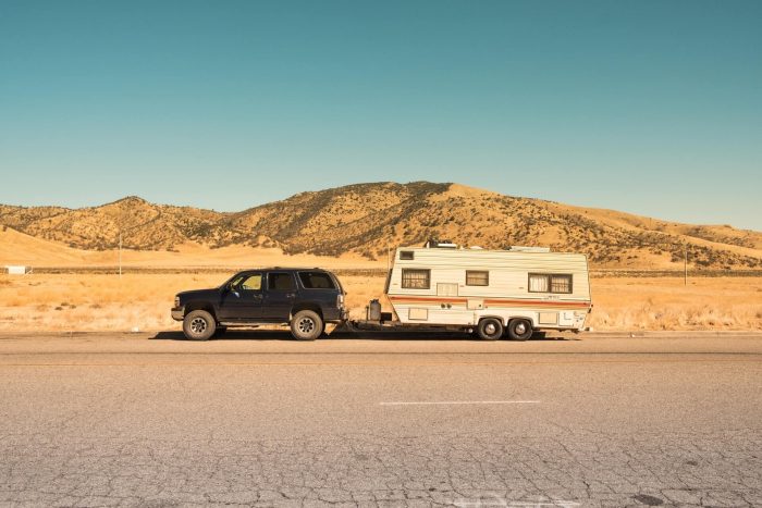 A photograph of a parked black SUV connected to a small white and orange caravan. Behind the caravan is a large desert and mountain range