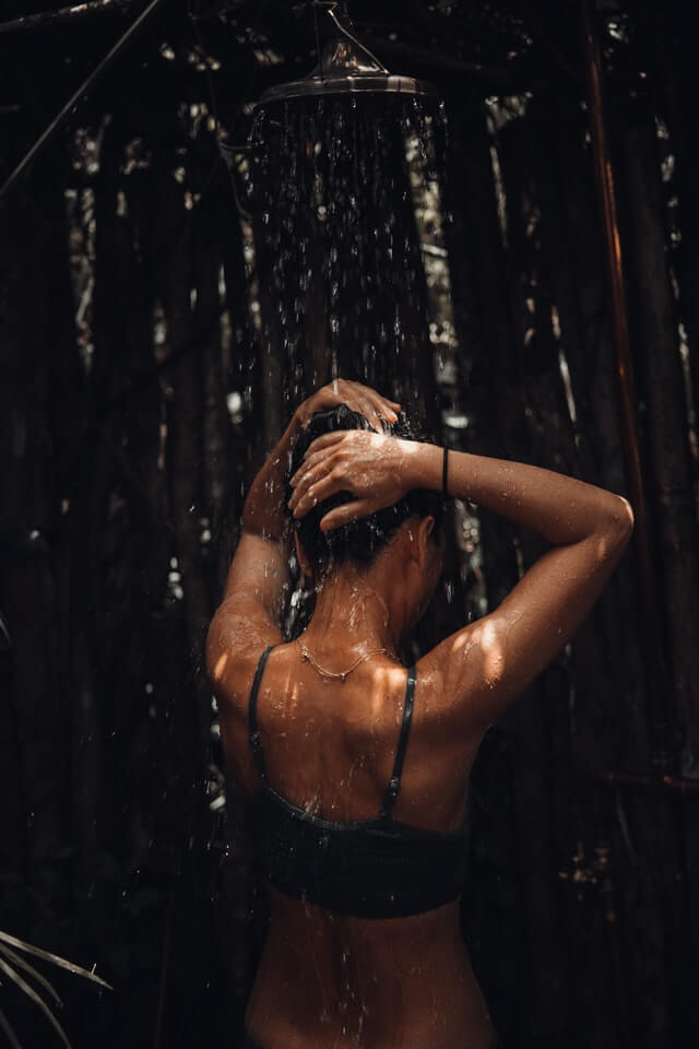 A photograph showing a woman standing in a shower, washing her hair. It shows the back of the woman.