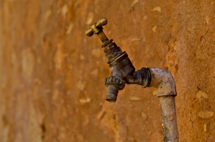 A picture of a rusty brass tap which has been damaged by acidic water. It shows an old outdoor tap in front of a brown wall